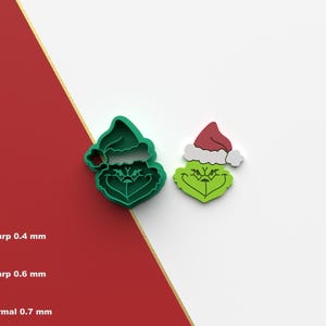 May include: Two green and red 3D printed cookie cutters shaped like the Grinch wearing a Santa hat. The Grinch is wearing a red Santa hat with a white trim and a green and white fur collar. The Grinch's face is green with a white beard and a grumpy expression.