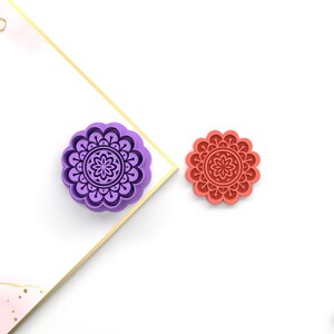 Puede incluir: Dos cortadores de galletas, uno morado y otro rojo, con forma de mandala con una flor en el centro.