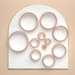 May include: A minimalist design featuring a white, arched panel with a gold border. Various-sized wooden rings are arranged on the panel, creating a layered, circular pattern. The background is a solid, light peach color, providing a soft contrast to the design.