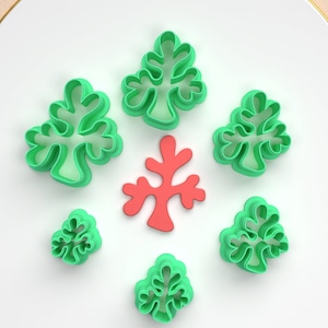 Puede incluir: Una colección de cortadores de galletas verdes con forma de árbol y un cortador rojo con forma de coral. Los cortadores están dispuestos sobre una superficie blanca, con un fondo suave y curvo.