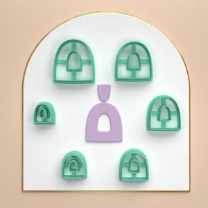 Puede incluir: Un conjunto de siete cortadores de galletas verdes y morados en forma de arcos y un solo arco. Los cortadores de galletas están dispuestos sobre una superficie blanca con un borde dorado.