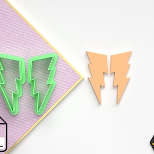 Può includere: Due tagliabiscotti a forma di fulmine in plastica verde su uno sfondo rosa e bianco. I tagliabiscotti sono accanto a due forme di fulmine arancioni.