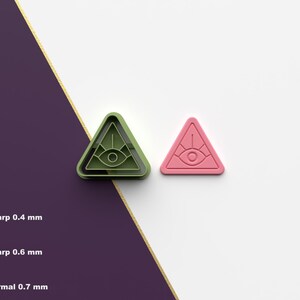 Puede incluir: Dos cortadores de galletas impresos en 3D en forma de triángulo con un ojo en su interior. Un cortador es verde y el otro es rosa.