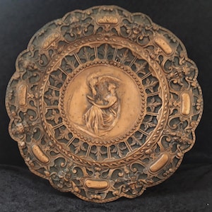 May include: A circular copper wall plaque with an ornate design. The center features a raised relief of a woman holding a bird. The plaque has a dark green patina.