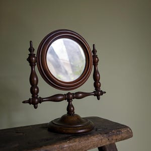 May include: A round tabletop mirror with a dark brown wooden frame and stand. The mirror is held by two vertical supports with decorative finials and a horizontal crossbar, all mounted on a circular base. The wood has a smooth, polished finish.