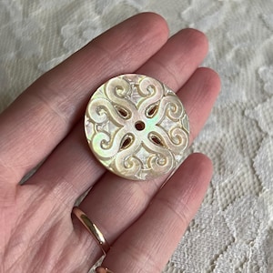 May include: A round, iridescent button with an ornate, swirling design. The button features a central hole and is made of a pearlescent material, reflecting light with a subtle sheen. The button's design is symmetrical and detailed.