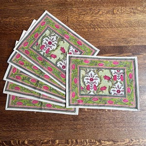 May include: A set of rectangular placemats with a floral design. Each placemat features a white border, a green and pink floral pattern, and a gray border. The placemats are arranged on a wooden surface.
