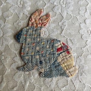 May include: A fabric bunny applique in a patchwork design. The bunny features a mix of patterned fabrics in blue, pink, red, and yellow. The bunny is set against a white lace background.