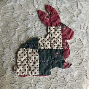 May include: A patchwork bunny applique in shades of red, green, and cream. The bunny is made of fabric squares with floral and heart patterns. The applique is placed on a cream-colored lace background, creating a vintage, handmade aesthetic.