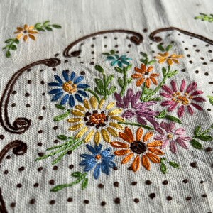 May include: A white linen tablecloth with a brown embroidered design of a bouquet of flowers. The flowers are in shades of yellow, orange, pink, blue, and green.