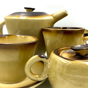 Op de afbeelding: Een set van bruine en beige keramische theekopjes, theepot, roomkan en suikerpot. De set heeft een rustieke, vintage uitstraling.
