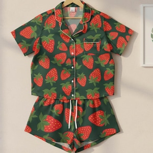 May include: A two-piece pyjama set featuring a dark green background and a pattern of red strawberries. The short-sleeved top and shorts are hanging on a wooden hanger. The set has white trim and a white drawstring.