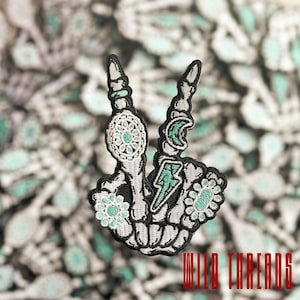 May include: An embroidered iron-on patch featuring a skeleton hand giving the peace sign. The hand is decorated with turquoise and white floral designs, a crescent moon, and a lightning bolt.