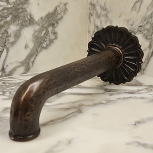 May include: A bronze-toned, curved door handle with a decorative floral rosette. The handle has a textured surface and is mounted on a white and gray marble surface. The handle's design suggests a vintage or antique style.
