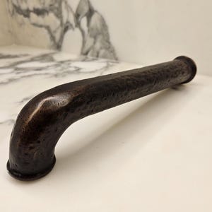 May include: A dark bronze-colored, curved metal handle with a textured surface. The handle has a cylindrical shape with a slight bend and is designed for pulling or gripping. The finish has a rustic, aged appearance.