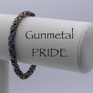 May include: Gunmetal bracelet with a rainbow colored chainmail design. The bracelet is displayed on a white fabric covered stand with the words "Gunmetal Pride" printed on it.