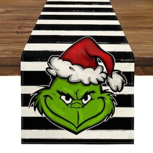 May include: A Christmas-themed table runner featuring a green Grinch face wearing a red Santa hat with white trim. The Grinch is set against a black and white striped background. The runner is rectangular and made of fabric.