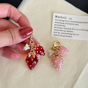 May include: Two strawberry-shaped charms with gold-colored accents. One charm features red strawberries with white dots, the other has pink strawberries. Each charm has small white flowers and pearls. The word "Musical" is printed on a card.