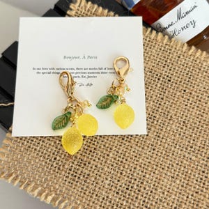 Puede incluir: Pendientes con colgantes dorados con encantos en forma de limón amarillo y detalles de hojas verdes. Los pendientes cuelgan de una tarjeta blanca con el texto "Bonjour, À Paris". Un tarro de miel está en el fondo.