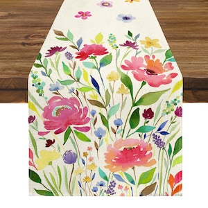 Puede incluir: Un camino de mesa rectangular con un diseño floral en acuarela. El camino de mesa presenta un fondo crema con una vibrante variedad de flores rosas, rojas, amarillas, moradas y azules y hojas verdes. El diseño está impreso en una tela texturizada.