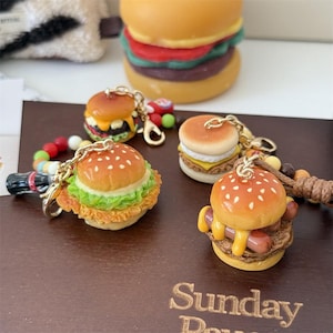 May include: A collection of miniature burger keychains with various toppings, including lettuce, cheese, and a sausage. The keychains have a golden chain and are set on a brown surface. A larger burger is in the background.