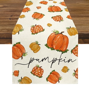 Peut inclure: Chemin de table crème avec un motif répété de citrouilles orange et jaunes avec des feuilles vertes. Le mot "pumpkin" est écrit en noir. Le chemin de table est posé sur une table en bois.