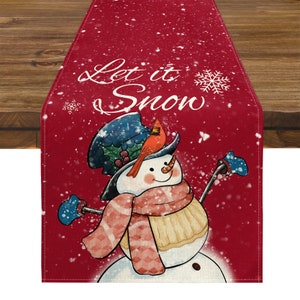 Op de afbeelding: Een rode kersttafelloper met een sneeuwmanontwerp en de tekst "Let it Snow". De sneeuwman draagt een blauwe hoed, een rode sjaal en blauwe wanten. Een rode kardinaal zit op de hoed van de sneeuwman. Sneeuwvlokken zijn verspreid.
