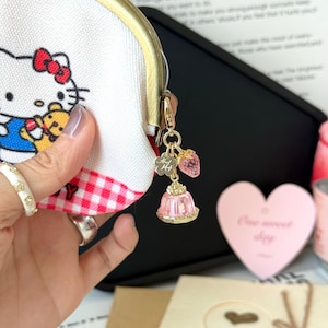 May include: A white coin purse with a gold clasp and a Hello Kitty design. The purse has a red and white gingham pattern at the bottom. A gold charm with a pink strawberry and a pink dessert charm hangs from the zipper.