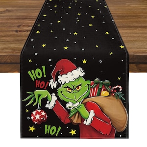 May include: Black Christmas table runner featuring a cartoon Grinch character in a Santa hat, holding a star ornament and a sack of gifts. The words "HO! HO!" are printed on the runner, along with yellow stars.