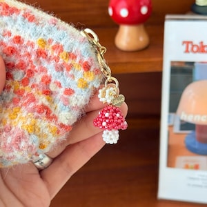Pode incluir: Uma pequena bolsa colorida com um fecho dourado e um berloque de cogumelo. O berloque é vermelho com pintas brancas e um caule branco. A bolsa tem um padrão floral em tons de vermelho, amarelo e azul.