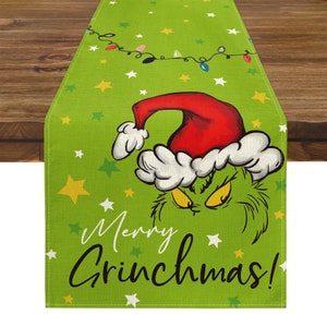 May include: Green Christmas table runner featuring the Grinch wearing a Santa hat. The runner has the text "Merry Grinchmas!" with star and string light accents. The table runner is made of a textured fabric.