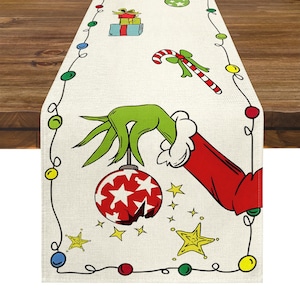 May include: A Christmas-themed table runner with a playful design. It features a green hand holding a red bauble with white stars, a candy cane, and gift boxes. The border is decorated with colourful lights and stars.