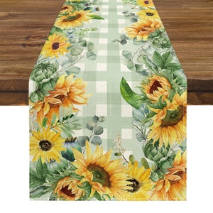 Puede incluir: Un camino de mesa rectangular con un patrón de cuadros verde claro y blanco. El camino de mesa está decorado con girasoles amarillos y follaje verde en los bordes. Los girasoles tienen pétalos amarillos y centros marrones.