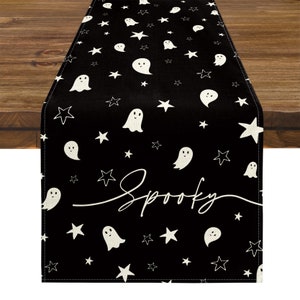 May include: Black Halloween table runner with white ghost and star patterns. The word "Spooky" is written in a cursive font across the center. The runner is placed on a wooden table.