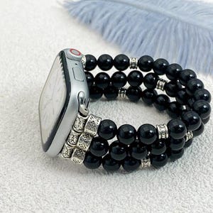 May include: Silver smartwatch with a black beaded bracelet. The bracelet has three strands of polished, round black beads, separated by silver-toned spacers. The watch case is silver with a red accent on the crown.