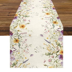 May include: Cream-coloured table runner with a floral design. The runner features a border of watercolour flowers in shades of yellow, purple, pink, and green. The flowers are arranged on a cream-coloured background, creating a spring or summer theme.