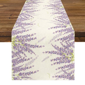 Pode incluir: Caminho de mesa retangular com um desenho floral de lavanda. O caminho de mesa tem um fundo creme com raminhos de lavanda roxos e folhas verdes. O caminho de mesa é colocado sobre uma mesa de madeira.