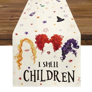 Może przedstawiać: Kremowy bieżnik na stół na Halloween z akwarelowymi ilustracjami włosów trzech wiedźm i napisem "I SMELL CHILDREN" w kolorze czarnym. Projekt zawiera gwiazdy, kapelusz czarownicy i butelki z eliksirami.