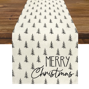 May include: A cream-colored Christmas table runner with a repeating pattern of black pine trees. The words "MERRY Christmas" are printed in black script and block letters. The runner is displayed on a wooden table.