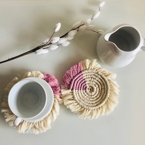 Könnte beinhalten: Zwei handgefertigte Makramee-Untersetzer in einem rosa-weißen Farbschema. Die Untersetzer haben einen Fransenrand und sind perfekt, um Oberflächen vor Hitze und Kratzern zu schützen.