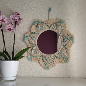 Può includere: Uno specchio rotondo con una cornice in macramé beige e blu chiaro. La cornice ha la forma di un fiore con frange attorno ai bordi.