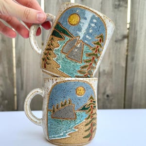May include: Two ceramic mugs with a landscape design. The mugs feature a blue sky with a yellow sun, a teal lake, and green and brown trees. The mugs are speckled with a tan base color and have a curved handle.