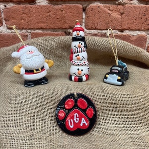 May include: Four ceramic Christmas ornaments: a Santa Claus, a snowman stack, a car, and a red paw print with the letters "UGA" in white.