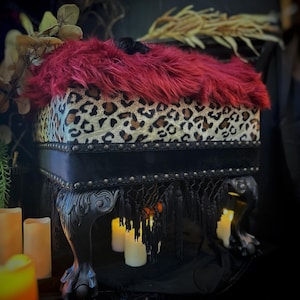 May include: A decorative footstool with a red faux fur top and a leopard print seat. The black frame features ornate claw feet and a black fringe. Candles are placed around the base, creating a gothic aesthetic.
