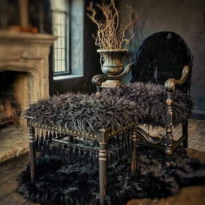 May include: A black armchair and matching ottoman set featuring long, shaggy faux fur upholstery. The wooden frame has a distressed finish with gold-tone nailhead trim and a row of hanging black tassels along the base of the ottoman.