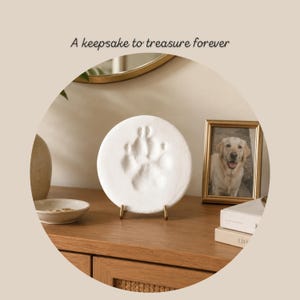 May include: A white, circular pet paw print keepsake on a gold stand. The image includes a framed photo of a golden retriever and the text "A keepsake to treasure forever."