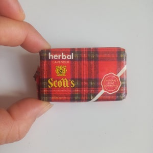 Pode incluir: Uma barra de sabonete vermelha e preta xadrez com as palavras "herbal lavender" e "Scott's" em letras douradas. O sabonete também tem um selo vermelho com as palavras "natural soap guarantee".