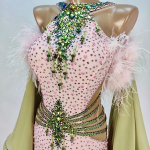 Puede incluir: Un vestido de baile rosa con un diseño de cristal verde e iridiscente. El vestido tiene un escote halter y un corpiño ajustado. La falda es larga y fluida. El vestido está adornado con plumas en los hombros.