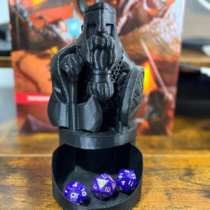 May include: A black 3D-printed dwarf figure with a long beard, helmet, and shield, atop a dice rolling tray. Three purple and white dice are inside the tray. The background includes a book with a fantasy theme.
