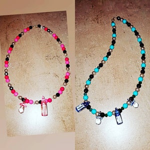 Può includere: Due collane con perline colorate e ciondoli. Una collana ha perline rosa, nere e argentate con un ciuccio rosa e un ciondolo a forma di biberon rosa. L'altra collana ha perline blu, nere e argentate con un ciuccio blu e un ciondolo a forma di biberon blu.
